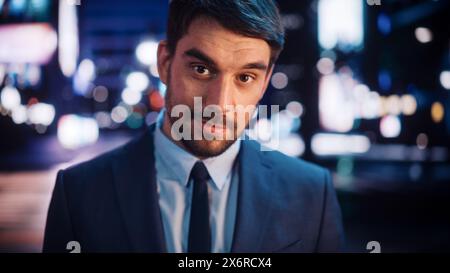 Ritratto dell'uomo barbuto sorridente, guardando la fotocamera, in piedi nella città notturna con bokeh Neon Street Lights sullo sfondo. Giovane uomo felice e sicuro di sé che indossa un abito elegante. Foto Stock