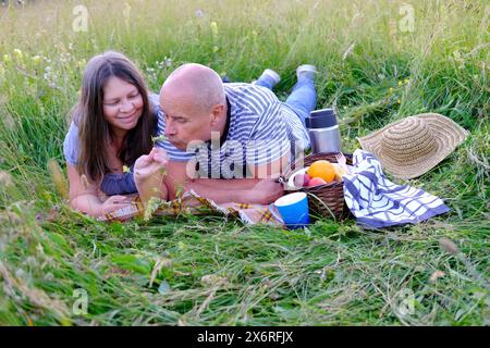 uomo maturo 60 anni, donna 50 anni in abiti blu giace sull'erba nel prato, accanto al cestino da picnic con frutta, thermos con tè, concetto di fam Foto Stock
