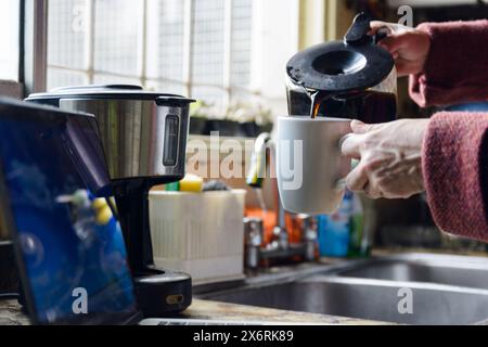 Il primo piano di una mano femminile irriconoscibile di una persona caucasica sta versando il caffè in una tazza da una macchina per il caffè in cucina vicino alla finestra del Foto Stock