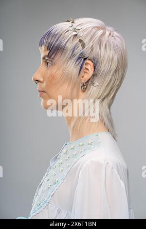 Ritratto laterale di una giovane donna - capelli biondi, evidenziazioni viola - perforazione del setto, orecchini multipli, top floreale trasparente, testa in foglia argento Foto Stock