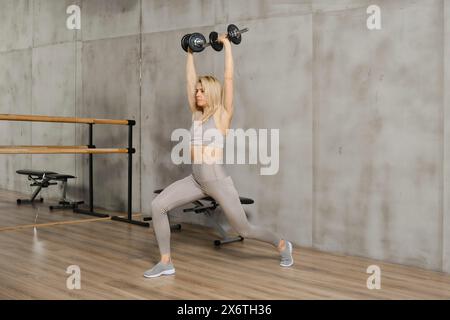 Donna di mezza età sportiva che fa affondamenti tenendo i manubri sopra la testa Foto Stock
