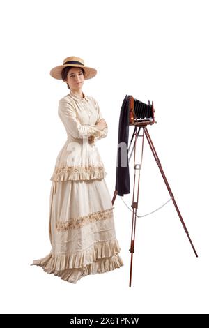 Giovane donna in abito vittoriano con un cappello di paglia in piedi accanto a una macchina fotografica su un treppiede Foto Stock