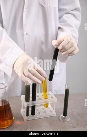 Donna che prende provette con diversi tipi di olio grezzo dal rack al tavolo grigio su sfondo chiaro, primo piano Foto Stock