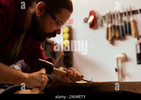 Scultore che dà forma al legno grezzo con scalpello e martello in falegnameria, crea arte del legno e indossa occhiali di sicurezza. Artista che realizza sculture in legno, incide legname con strumenti, utilizzando dispositivi di protezione Foto Stock