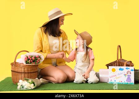 Giovane donna con la sua piccola figlia e cestini da picnic sull'erba su sfondo giallo. Festa della mamma Foto Stock