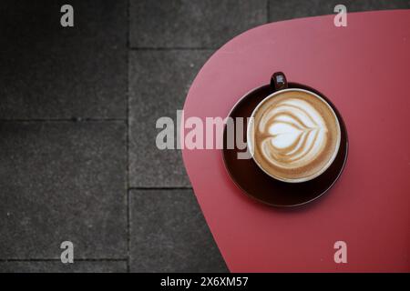 Un latte splendidamente realizzato con latte art a forma di cuore, servito in una tazza e un piattino marrone, collocato su un tavolo rosso. Foto Stock