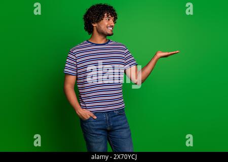 Foto ritratto di giovane studente indiano ragazzo divertente con capelli ondulati t shirt a righe tenendo a mano prezzo affare isolato su sfondo di colore verde Foto Stock