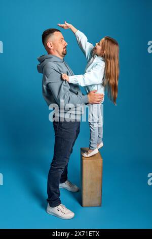 Il ritratto di una bambina in piedi e misura la sua altezza con suo padre, mostrando come è cresciuta su sfondo blu. Foto Stock