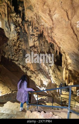 Una donna sta su un passaggio pedonale in una grotta, guardando verso il soffitto. La grotta è piena di stalattiti e stalagmiti, creando un senso di stupore e. Foto Stock