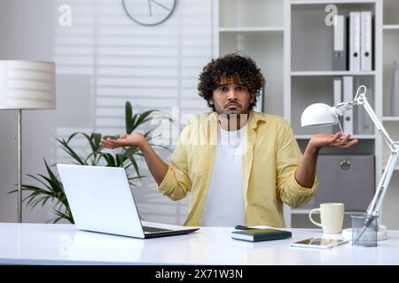Giovane che sembra confuso, che si stringe le spalle con incertezza mentre lavora su un notebook in un ufficio. Foto Stock