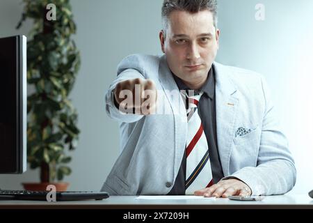 Uomo di mezza età con blazer grigio chiaro, camicia grigio scuro e cravatta a righe, con capelli grigi piccanti, estende il pugno in avanti, mostrando un look serio, il mare Foto Stock