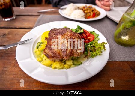 Succosa bistecca di maiale con insalata leggera di patate e verdure su piatto bianco, secondo piatto sullo sfondo con riso, carne di pollo con pepe rosso, bevanda, coltello Foto Stock