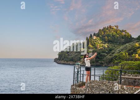 Adolescente (13 anni) che fa un selfie con il suo smarphone sulla Terrazza di Punta Caiega, con il promontorio di Portofino, Genova, Liguria, Italia Foto Stock