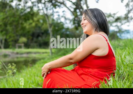 donna latina in abito rosso seduta sul prato, guardando verso l'orizzonte sorridendo Foto Stock