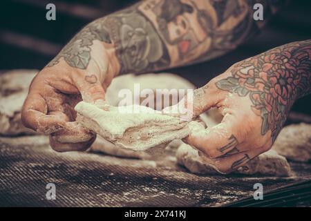 Un fornaio con tatuaggi impasta l'impasto del pane nella panetteria. Stile vintage con grana. Foto Stock
