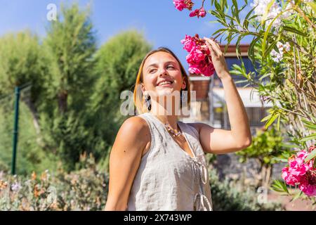 Una donna sta in piedi in un giardino, sorride e tiene in mano un fiore rosa. La scena è luminosa e allegra, con il sorriso della donna che riflette l'a positiva Foto Stock