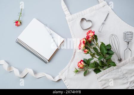 Taccuino vuoto aperto con fiori rose, accessori da forno sul grembiule su sfondo grigio, piatto, vista dall'alto. Ricette di cottura festive Foto Stock