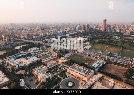 Vista aerea del Cairo che mostra un ponte sul Nilo, un fitto paesaggio urbano, campi sportivi, piscine ed edifici governativi sotto la luce soffusa del tardo pomeriggio. Foto Stock