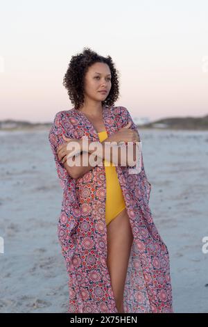 In spiaggia, giovane donna birazziale in piedi da sola, che attraversa le braccia Foto Stock