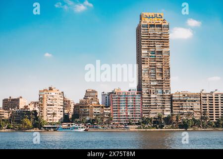 Il lungofiume del Nilo al Cairo presenta alte torri residenziali, una lunga barca turistica e un imponente grattacielo con un cartello arabo giallo Foto Stock