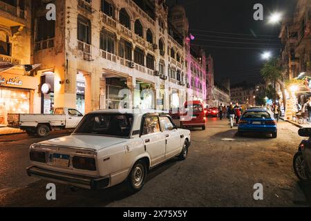 Scena di strada notturna a Heliopolis, il Cairo, con una Lada d'epoca parcheggiata accanto all'architettura storica, al traffico vivace e alle luci calde del punto vendita Foto Stock