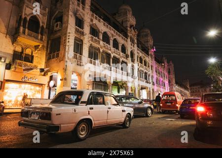 Scena di strada notturna a Heliopolis, il Cairo, con una Lada d'epoca parcheggiata accanto all'architettura storica, al traffico vivace e alle luci calde del punto vendita Foto Stock