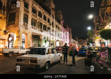 Scena di strada notturna a Heliopolis, il Cairo, con una Lada d'epoca parcheggiata accanto all'architettura storica, al traffico vivace e alle luci calde del punto vendita Foto Stock