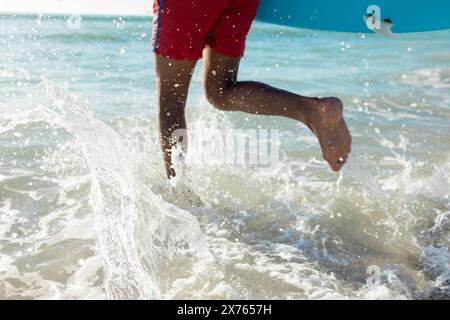 In spiaggia, giovane birazziale che tiene la tavola da surf e corre in mare Foto Stock