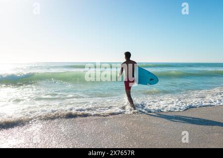 In spiaggia, giovane uomo birazziale che tiene la tavola da surf che corre verso il mare Foto Stock