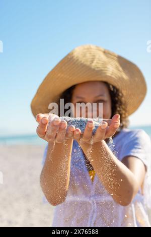 In spiaggia, giovane donna birazziale che indossa un cappello da sole che regge la sabbia Foto Stock