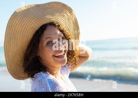 In spiaggia, giovane donna birazziale che indossa un cappello a teglia larga sorridente Foto Stock
