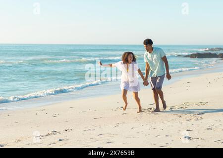 In spiaggia, coppia birazziale che si tiene per mano, cammina, entrambi sorridenti Foto Stock