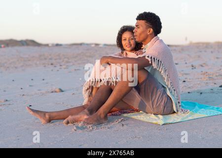 In spiaggia, coppia birazziale seduta, avvolta in coperta, che guarda l'orizzonte Foto Stock
