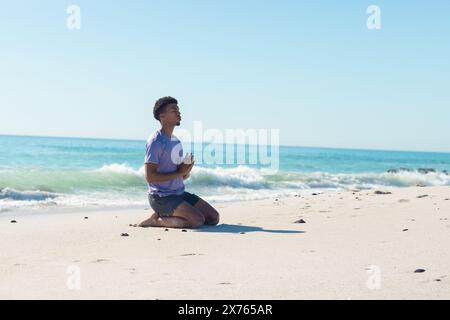 In spiaggia, giovane birazziale seduto, mani unite, occhi che si chiudono Foto Stock