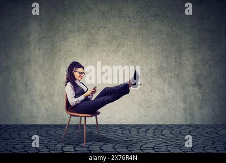 Giovane donna d'affari felice seduta su una sedia con le gambe sollevate, che chiacchiera sul suo telefono cellulare Foto Stock