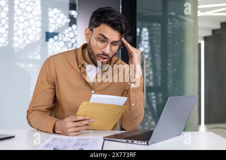 Un uomo scioccato legge una lettera con cattive notizie in ufficio mentre lavora sul suo portatile. Preoccupazione e stress per gli affari. Foto Stock