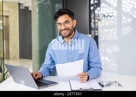 Uomo d'affari sorridente in camicia blu che lavora su un computer portatile e rivede documenti alla scrivania con tablet e documenti. Foto Stock