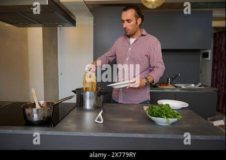 Un giovane chef maschile che riversa il passato italiano appena cucinato in un piatto, pronto a servirlo ai clienti. Consumismo di cibo e bevande. Epicura. Cu Foto Stock