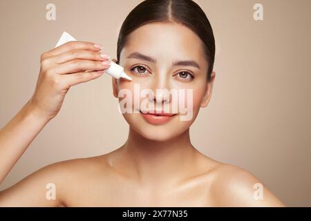 Bella donna che usa la crema intorno agli occhi. Video di donna con trucco perfetto su sfondo beige. Concetto di bellezza e cura della pelle Foto Stock