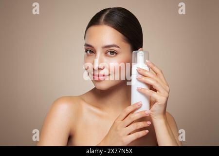 Una giovane donna mostra una bottiglia con un dispenser di schiuma per il lavaggio. Pubblicità quotidiana di cosmetici per la cura della pelle. Studio Shoot Beige background Foto Stock