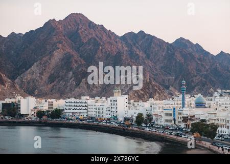 Vista panoramica della Mutrah Corniche a Mascate, Oman. Edifici bianchi, moschea con cupola blu e porto adagiato contro le spettacolari montagne rocciose al crepuscolo Foto Stock