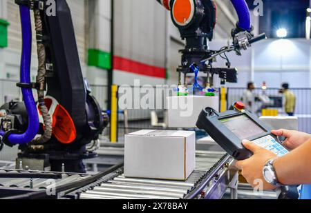 Il responsabile controlla e controlla i bracci del robot di automazione che tengono una scatola bianca e il lavoratore che aziona una macchina robotizzata con un pannello di controllo Foto Stock