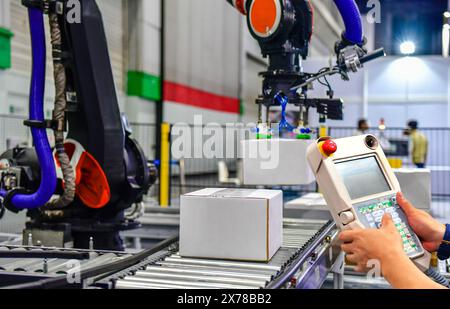 Il responsabile controlla e controlla i bracci del robot di automazione che tengono una scatola bianca e il lavoratore che aziona una macchina robotizzata con un pannello di controllo Foto Stock