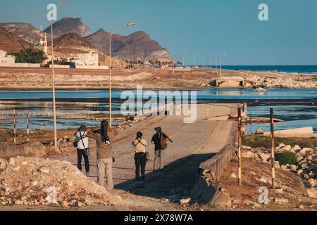 Gli osservatori di uccelli scattano foto al ponte rotto presso al Mughsail Beach in Oman, con montagne aspre e mare sullo sfondo. Foto Stock