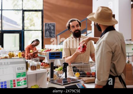 Un giovane venditore che indossa un grembiule e un cappello aiuta i clienti caucasici ad acquistare cibi freschi e rispettosi dell'ambiente in un negozio nelle vicinanze. Varie opzioni alimentari nutrienti e imballaggi riciclabili. Foto Stock