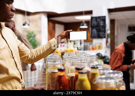 In un negozio di alimentari ecologico, il consumatore afroamericano di sesso maschile ha un cellulare che mostra uno schermo bianco isolato. L'immagine mostra un uomo nero con dispositivo mobile dotato di un modello cromakey. Foto Stock