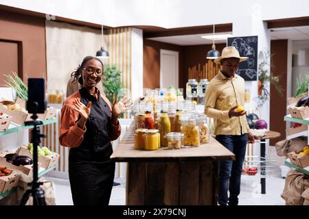 Il proprietario di un'azienda afroamericana filma vlog che promuovono prodotti freschi e biologici in un negozio ecologico, utilizzando smartphone e bastone per selfie. L'immagine mostra una donna nera che parla davanti al dispositivo mobile. Foto Stock
