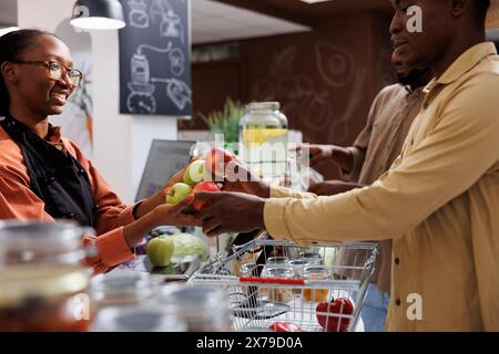 Il proprietario afroamericano aiuta i clienti in un negozio ecologico con prodotti biologici freschi. Attenzione selettiva su un cliente maschio che consegna frutta alla cassiera femminile al banco cassa. immagine vista laterale. Foto Stock