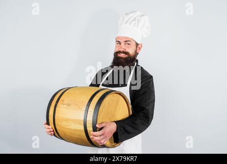 Birraio maschio in grembiule che tiene la botte con birra artigianale presso la fabbrica di birra. Uomo barbuto in cappello da chef e uniforme con fusto di legno. Birra tradizionale tedesca Foto Stock