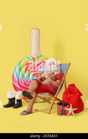 Babbo Natale che suona ukulele su una sedia a sdraio su sfondo giallo. Natale a luglio Foto Stock